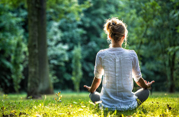 Vrouw in wit shirt zit te mediteren op een grasveld in de zon met haar rug naar de kijker toe en haar handpalmen open op haar knieën