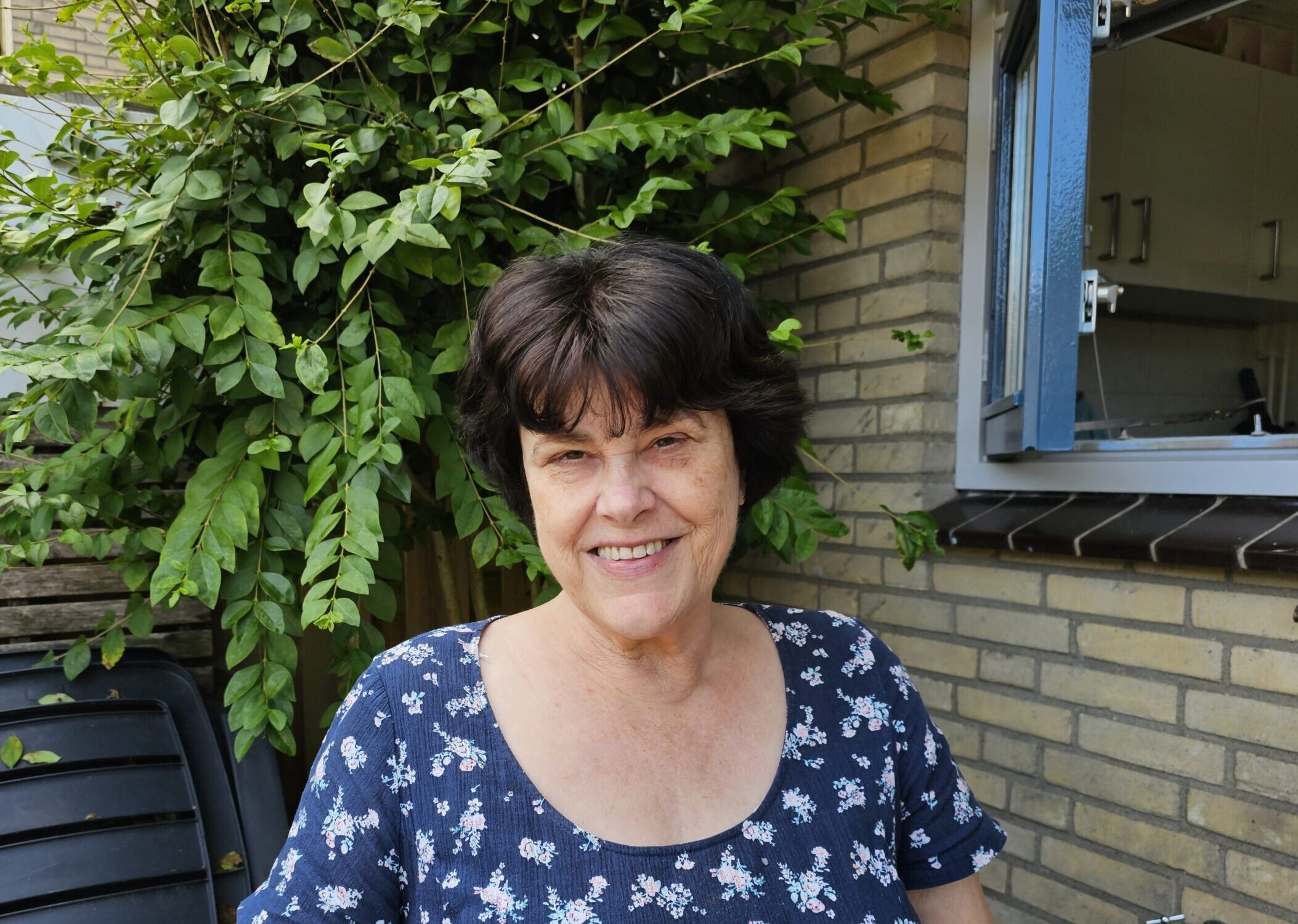 Vrouw met kort donker haar en pony, lachend gezicht en donkerblauwe bloemetjesjurk in de tuin bij open keukenraam. NAH en heupoperaties